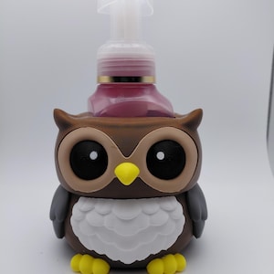 May include: A brown and gray owl-shaped soap dispenser. The dispenser has large black eyes, a yellow beak, and white chest feathers. The soap bottle inside is pink, and the pump is clear. The owl's feet are yellow.