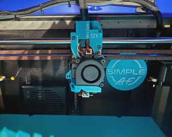 May include: A close-up of a 3D printer's print head assembly. The assembly is primarily teal, with a circular fan and the text "SIMPLE AF" on a teal circle. The printer's frame is visible in the background.