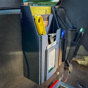 May include: A black, textured organiser with multiple compartments. A yellow tool and paperwork are visible inside. A clipboard with a card is attached to the front. A black pouch with pens is to the right.
