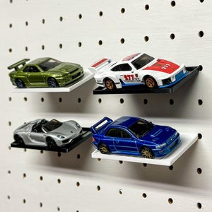 Pode incluir: Quatro carros de brinquedo expostos em prateleiras brancas com um fundo de placa de pinos. Os carros são um Nissan Skyline verde, um Porsche 911 branco, um Porsche 918 Spyder prateado e um Honda Civic Type R azul.