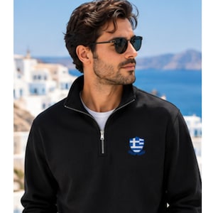 Puede incluir: Una sudadera negra con cremallera de un cuarto de largo y un pequeño escudo bordado en el pecho izquierdo. El emblema muestra el patrón de rayas horizontales azules y blancas de la bandera griega. La prenda tiene mangas largas y una cremallera plateada.
