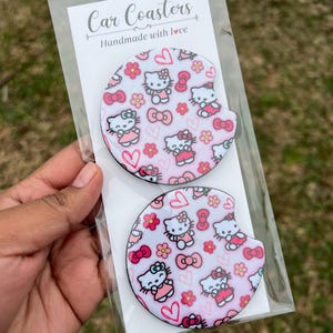 May include: Two round car coasters with a white background and a pink and white Hello Kitty pattern. The coasters are packaged in a clear plastic bag with a white label that reads "Car Coasters Handmade with Love".