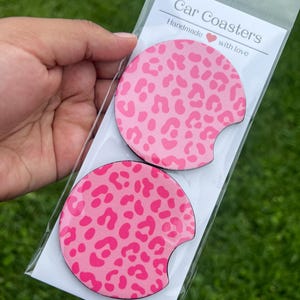 May include: Two pink car coasters with a leopard print design. The coasters are round with a curved cutout for easy removal. The packaging reads "Car Coasters, Handmade with love."