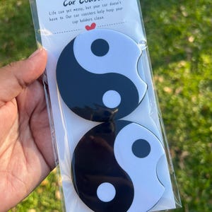 May include: Two car coasters with a yin-yang design, black and white, are packaged in a clear plastic sleeve. The text "Car Coaster" and a short description are printed on the packaging.