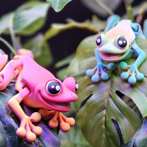 May include: Two whimsical frog figurines in vibrant colors rest on large green leaves. One frog is pink with orange accents, the other is light green and blue. Both have large, expressive eyes and are set against a dark background.