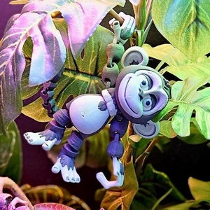 May include: Three small monkey figurines are posed on a bed of green and purple foliage. The monkeys are gray and have black eyes. The monkey in the center is holding a small, brown branch. The monkey on the left is sitting on a rock. The monkey on the right is hanging from a branch.