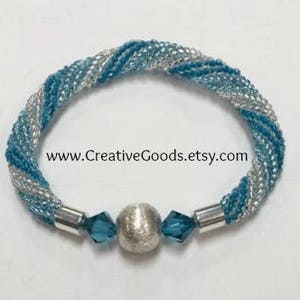 May include: A twisted beaded bracelet with silver and blue beads. The bracelet has a silver bead in the center with two blue crystal beads on either side. The bracelet is on a white background. www.CreativeGoods.etsy.com