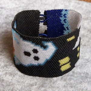 May include: A black beaded bracelet with a white and blue design. The bracelet features a pattern of ghosts with black eyes and a yellow stripe.
