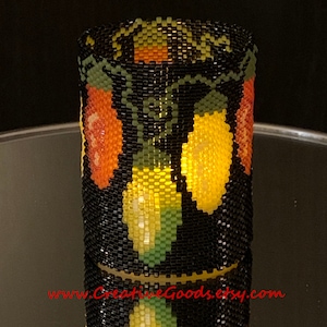 Puede incluir: Funda cilíndrica de cuentas con fondo negro, con un patrón de luces de colores en naranja, amarillo y verde. La funda está probablemente diseñada para encajar alrededor de un vaso u objeto similar. La dirección del sitio web es visible en la parte inferior.