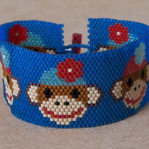 Puede incluir: Un brazalete de cuentas azules con un patrón repetido de monos que llevan sombreros rojos y flores azules. Los monos tienen pelaje marrón, caras blancas y labios rojos.