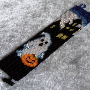 May include: A black beaded bracelet with a white ghost holding a jack-o'-lantern, a black house with yellow windows, and a blue sky with white stars.