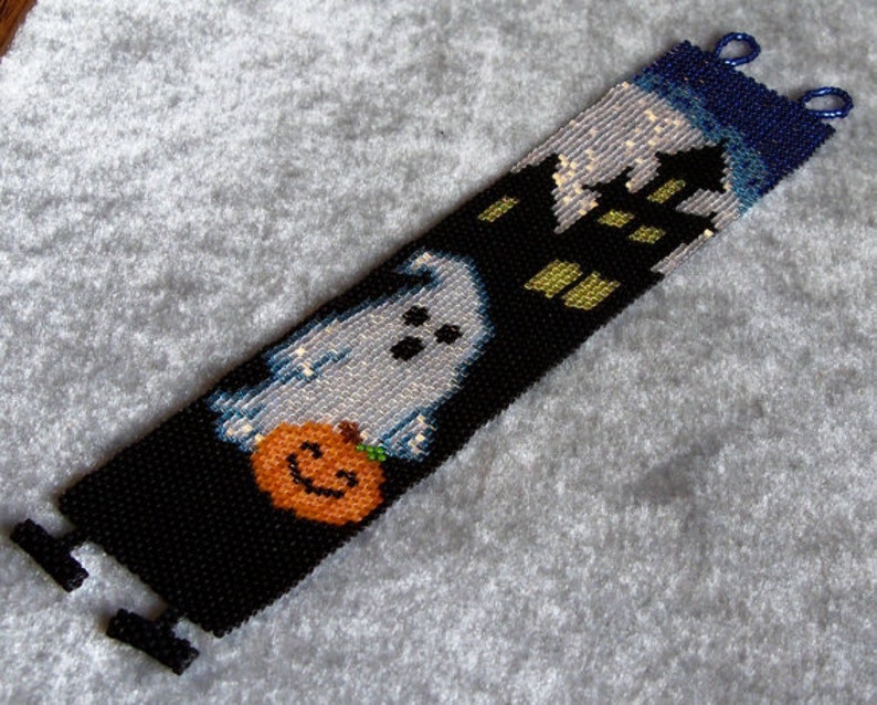 May include: A black beaded bracelet with a white ghost holding a jack-o'-lantern, a black house with yellow windows, and a blue sky with white stars.