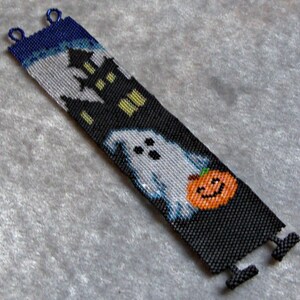 Op de afbeelding: Een zwart-wit kralenarmband met een spook, een huis en een pompoen ontwerp. De armband heeft blauwe en zilveren kralen.