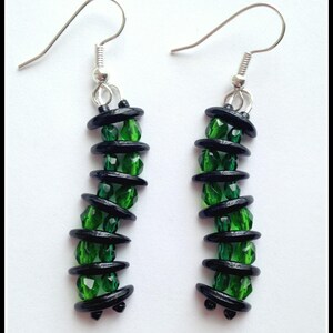 May include: A pair of dangle earrings featuring a combination of green faceted beads and black, oval-shaped beads. The earrings have silver-colored ear wires and a vertical design, creating a striking contrast of colors and textures.