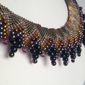 Op de afbeelding: Een veelkleurige kralenketting met een franje van zwarte kralen. De ketting is gemaakt met verschillende iriserende kralen in tinten goud, bruin, roze en paars.