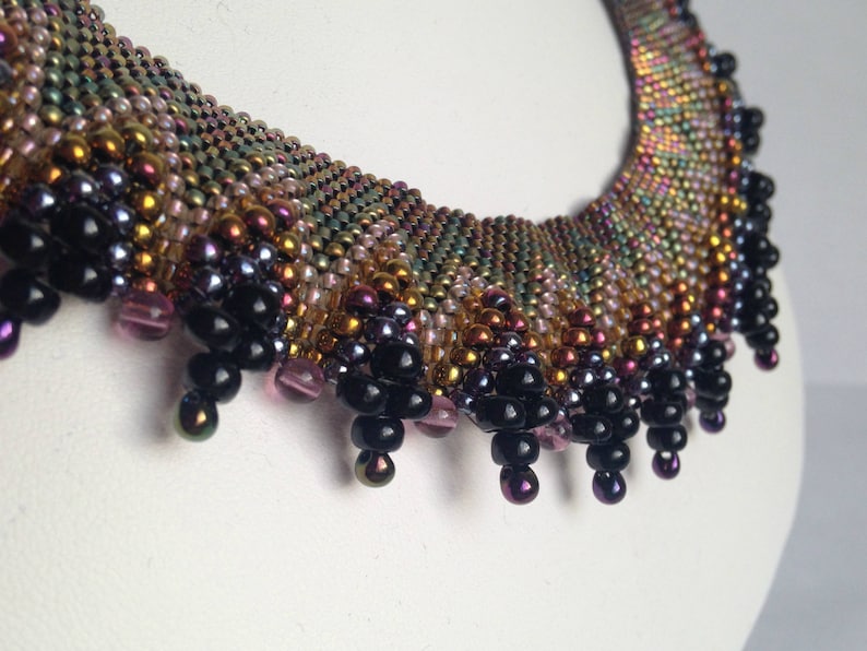 Op de afbeelding: Een veelkleurige kralenketting met een franje van zwarte kralen. De ketting is gemaakt met verschillende iriserende kralen in tinten goud, bruin, roze en paars.