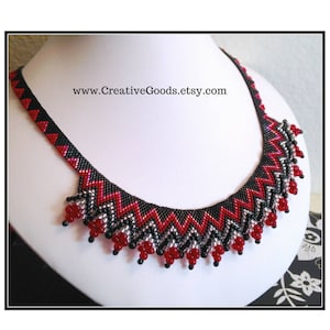Puede incluir: Un collar de cuentas negras, rojas y plateadas con un patrón geométrico. El collar tiene una franja de cuentas rojas en la parte inferior.