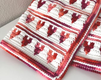 May include: A white crocheted blanket with a pink and red heart pattern. The blanket has a crocheted border in shades of pink and red.