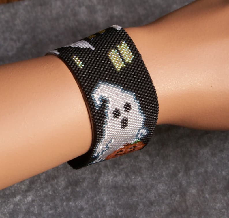 May include: A black beaded bracelet with a white ghost and orange pumpkin design. The bracelet is on a person's wrist.