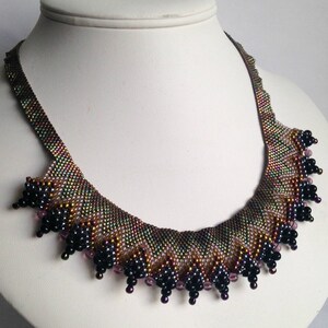 Op de afbeelding: Een kralenketting met een aflopende franje van zwarte en paarse kralen. De ketting is gemaakt van iriserende bruine, groene en gouden kralen.