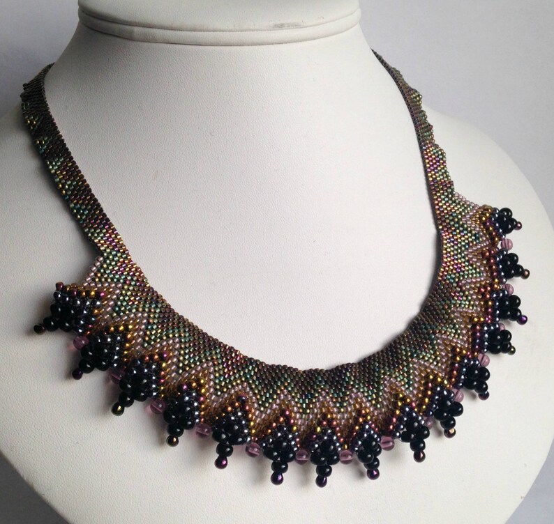 Op de afbeelding: Een kralenketting met een aflopende franje van zwarte en paarse kralen. De ketting is gemaakt van iriserende bruine, groene en gouden kralen.