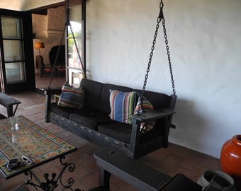 May include: A black wooden swing bench with striped cushions hangs from a chain in a patio setting. The swing is in front of a white wall and a tiled floor.