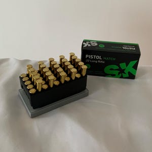 May include: A black and gray ammunition holder filled with gold-colored bullets. A box of .22 Long Rifle Pistol Match ammunition is next to the holder. The box is black with green and white text and a green 'SX' logo.