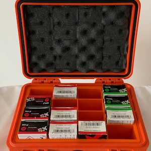May include: An open orange hard case with black foam padding and compartments holding small boxes. The boxes are labeled with text and barcodes, containing ammunition for rifles and pistols.