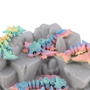 May include: Several articulated dragon figurines in pastel colours of blue, pink, yellow, and green. The dragons are arranged on a grey, textured rock formation, each with a rose-like detail. The figurines are a charming decorative item.
