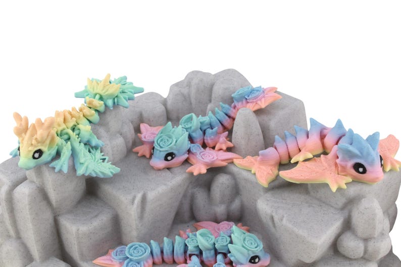 May include: Several articulated dragon figurines in pastel colours of blue, pink, yellow, and green. The dragons are arranged on a grey, textured rock formation, each with a rose-like detail. The figurines are a charming decorative item.