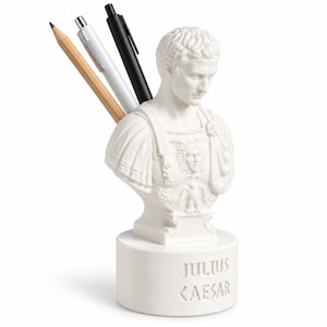 May include: A white desk organizer shaped like a bust of Julius Caesar, featuring a cylindrical base with the text JULIUS CAESAR embossed on the front. The sculpture holds a wooden pencil, a white pen, and a black pen in the back section.