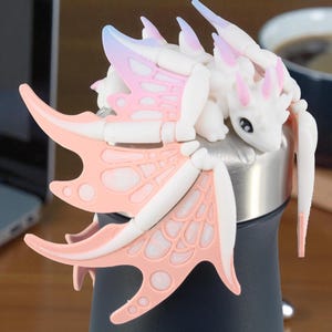May include: A white and pink dragon figurine with detailed wings and a whimsical design. The dragon is perched on a dark blue and silver object, possibly a bottle or container. The wings have a pink and white color scheme.