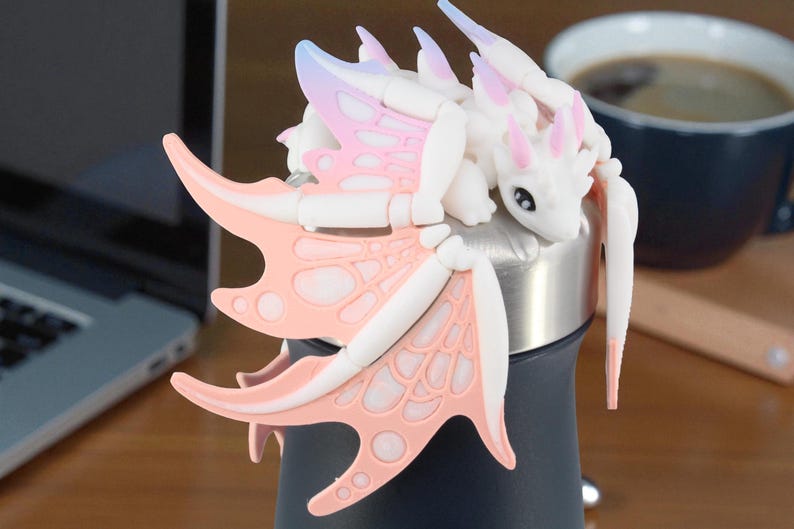 May include: A white and pink dragon figurine with detailed wings and a whimsical design. The dragon is perched on a dark blue and silver object, possibly a bottle or container. The wings have a pink and white color scheme.