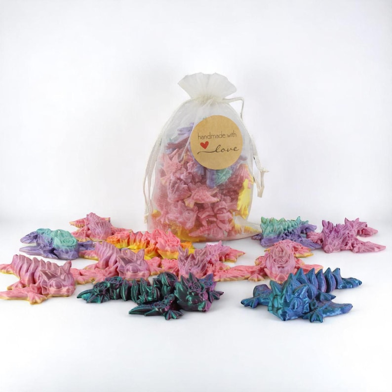 May include: A selection of multi-coloured, articulated dragon figurines in shades of pink, purple, green, and blue. Some dragons are scattered, while others are inside a sheer bag with a "handmade with love" tag.