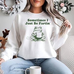 May include: White sweatshirt with a green frog graphic holding a mug, and the text "Sometimes I Just Be Fartin'". The frog is sitting with a puff of smoke. The sweatshirt is a casual, comfortable style.