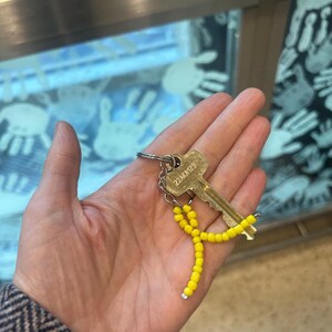 Puede incluir: Una llave dorada con un charm de cinta de cuentas amarillas. La llave tiene el texto "22AK12" en ella.