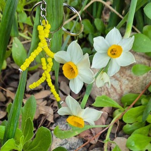Puede incluir: Un llavero de cuentas amarillas con forma de persona que cuelga de un llavero plateado. El llavero est&aacute; delante de narcisos blancos con centros amarillos.