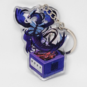 May include: A clear acrylic keychain featuring a stylized, dark blue dragon over a purple video game console. The dragon has a red eye and is surrounded by dark blue and black accents. A silver chain and keyring are attached.