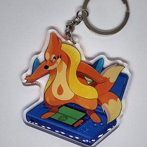 May include: A clear acrylic keychain featuring a cartoon fox-like character with orange and yellow fur, standing on a blue handheld game console. The keychain has a silver keyring and chain.