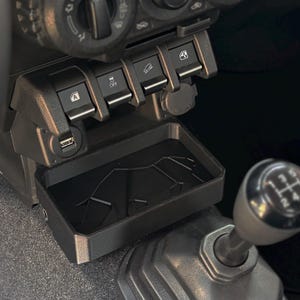 Puede incluir: Una bandeja de almacenamiento de plástico negro con una superficie texturizada se encuentra en la consola central de un automóvil. La bandeja está ubicada junto a un puerto USB y una toma de corriente de 12V. La bandeja está diseñada para contener objetos pequeños, como teléfonos, llaves y monedas.