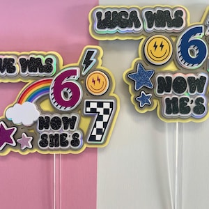 May include: Two birthday cake toppers with text and decorative elements. One says "Maeve was 6, now she's 7" with a rainbow and stars. The other says "Luca was 6, now he's 7" with lightning bolts and smiley faces.
