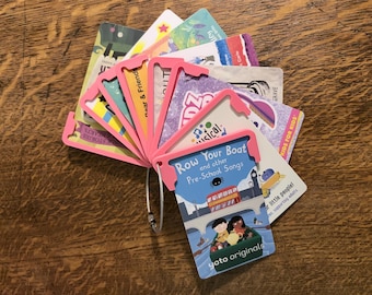 May include: A collection of rectangular Yoto audio cards held together by a silver metal ring and pink plastic protective frames. The top card features a colorful illustration of a boat on a river with the text Row Your Boat and other Pre-School Songs.