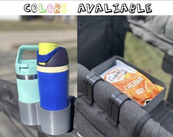 May include: Image features a baby stroller accessory set. Includes a dual cup holder with a teal and blue water bottle, and a snack tray with a bag of cheddar crackers. The text "COLORS AVAILABLE" is at the top.