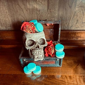 May include: A white ceramic skull with a blue and white design, filled with small blue containers. The skull is sitting in a wooden box with a red rose and other flowers.
