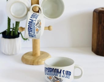 Puede incluir: Un soporte de madera para tazas con tres tazas blancas y un cuenco, con una cuarta taza delante. Las tazas y el cuenco tienen la inscripción "MUSHROOM SOUP" en azul, con un diseño de seta. Una pequeña planta está en el fondo.