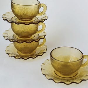 May include: A stack of three amber-colored glass teacups with matching saucers. The cups have a textured surface and small handles. The saucers have a scalloped edge. A fourth cup and saucer are separate.