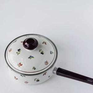 Puede incluir: Cacerola de esmalte blanco con tapa a juego. La cacerola y la tapa están decoradas con un estampado floral en tonos azules, rojos y amarillos. El asa es de color burdeos oscuro.