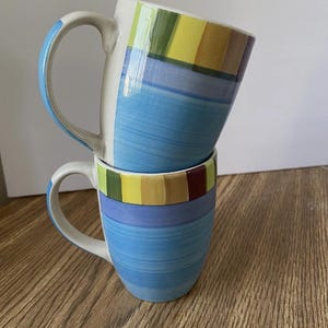 以下が含まれることがあります： 2つのセラミックマグカップが積み重ねられています。マグカップは青色で、水平ストライプと黄色、緑、赤のカラフルなバンドがあります。各マグカップには白いハンドルが付いています。