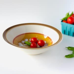 Puede incluir: Un cuenco de cerámica blanco con borde marrón y dorado, que contiene tres tomates cherry rojos y un pimiento verde. Una pequeña cesta azul de tomates está en el fondo. El cuenco tiene un diseño amarillo y rojo.