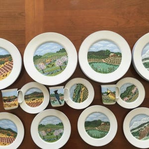 May include: A collection of white ceramic plates and mugs with landscape paintings. The plates vary in size and feature scenes of rolling hills, fields, and trees. The mugs also depict similar landscape scenes, with one showing a bird.